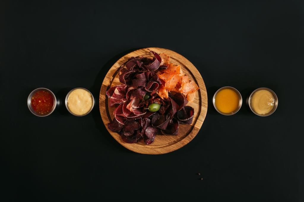 top view of delicious sliced assorted meat on wooden board with various sauces on black - Photo, Image