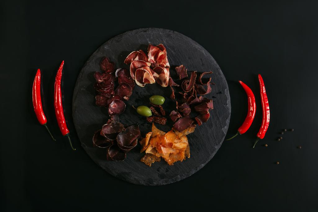 top view of gourmet assorted meat with olives on slate board and chili peppers on black - Photo, Image