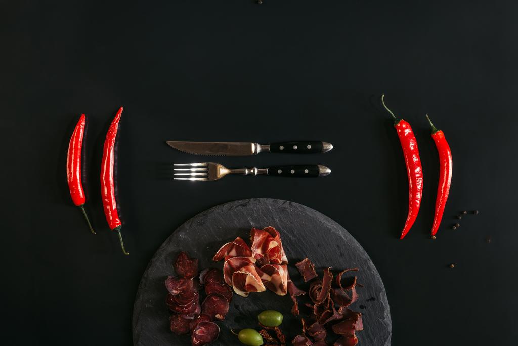 top view of gourmet assorted meat on slate board, fork with knife and chili peppers on black  - Photo, Image