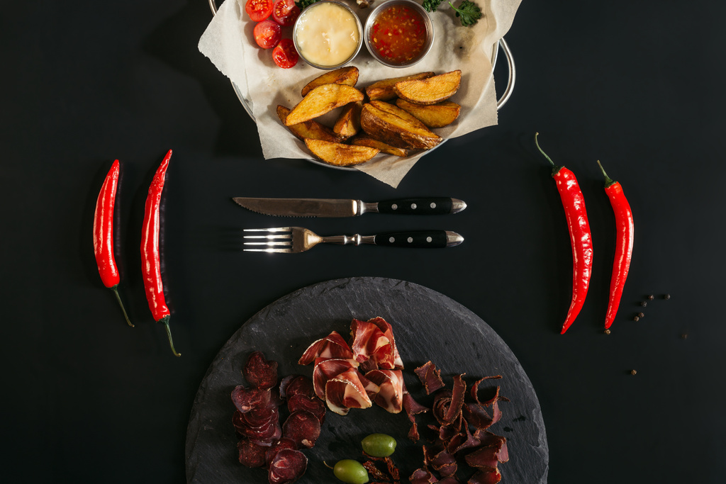 pohled shora na pečené brambory s omáčky, chilli papričky, vidličku, nůž a gurmánské míchané maso na břidlicové desky na černém pozadí - Fotografie, Obrázek