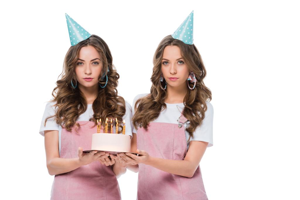 привлекательные молодые близнецы, держащие торт на день рождения и смотрящие на камеру, изолированную на белом
 - Фото, изображение