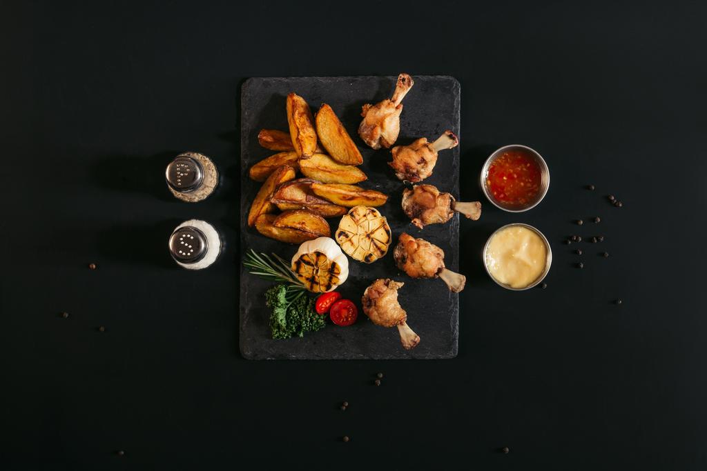 вкусный жареный картофель с курицей и чесноком на гриле на доске с приправами и соусами на черном
  - Фото, изображение