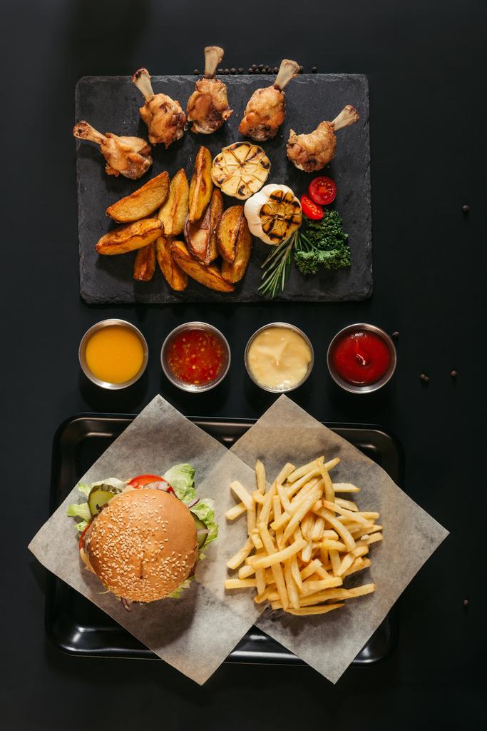 το Top view τηγανητές με νόστιμο burger στη θήκη, ανάμικτες σάλτσες και πλακών σκάφους με ψητές πατάτες και ψητά λαχανικά και φτερούγες κοτόπουλου σε μαύρο - Φωτογραφία, εικόνα