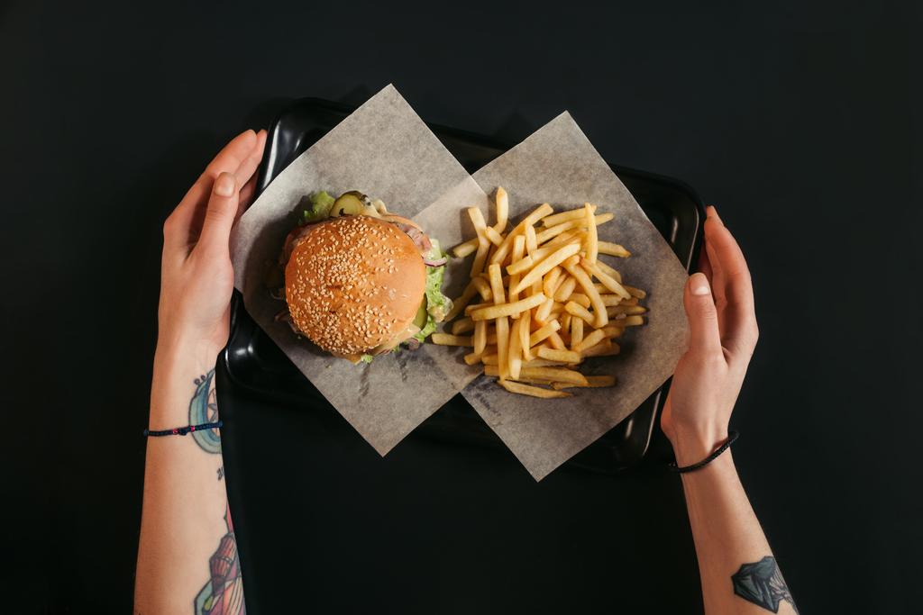 plan recadré de mains humaines tenant plateau avec hamburger délicieux et frites sur noir
 - Photo, image