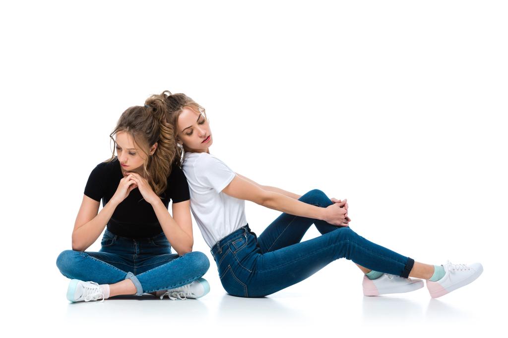 привлекательные молодые близнецы в джинсах и футболках, сидящие на полу на белом
 - Фото, изображение
