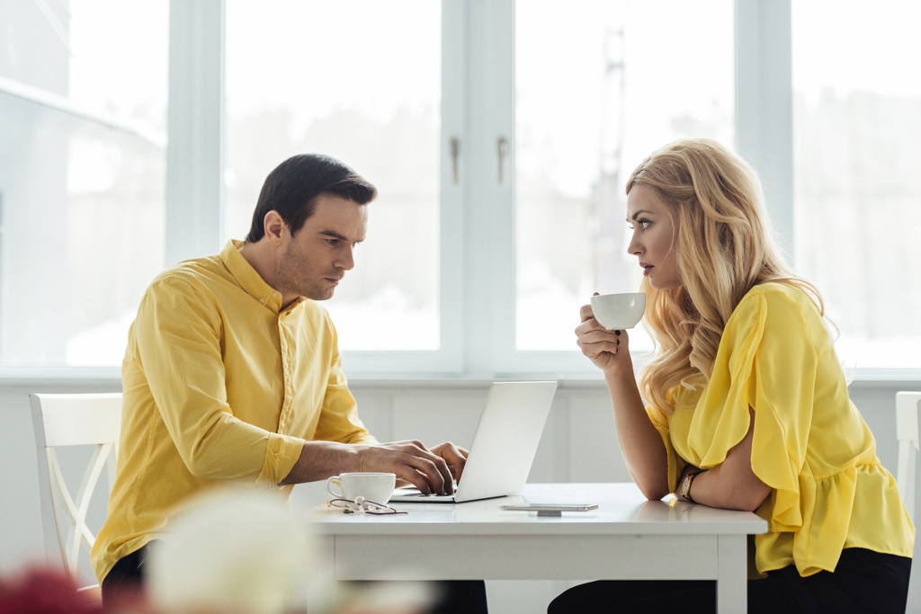 Blonde vrouw koffie drinken en kijken naar nadenkende man aan het werk op laptop - Foto, afbeelding