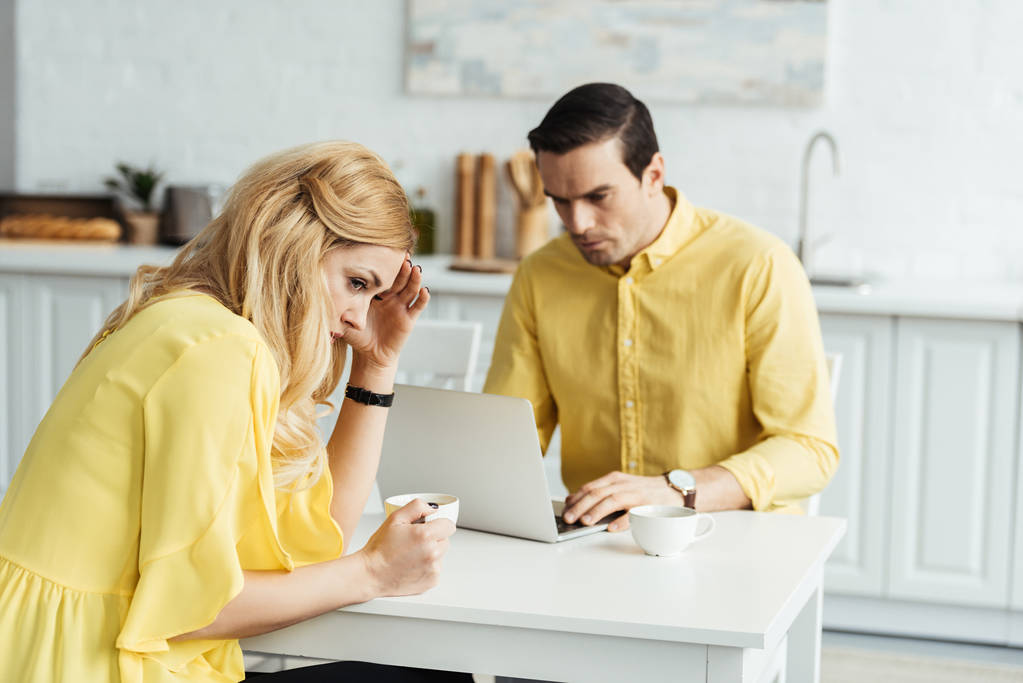 Upset woman with coffee sitting by man working on laptop in kitchen - Photo, Image