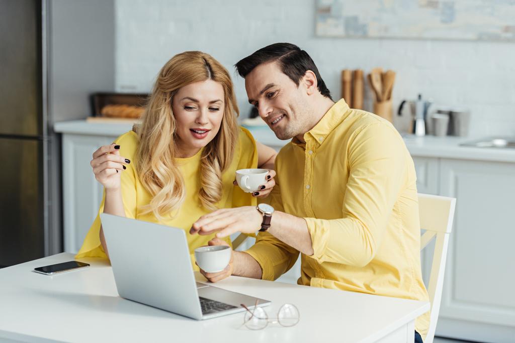 Чоловік і жінка п'ють каву і дивляться на екран ноутбука за кухонним столом
 - Фото, зображення