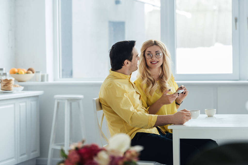 Man and woman with smartphone drinking coffee and talking by kitchen table - Photo, Image