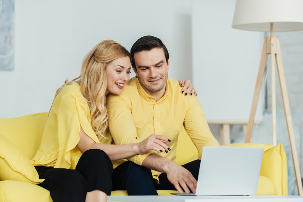 Ευτυχισμένο ζευγάρι κάθεται σε κίτρινο καναπέ και εργάζεται από το laptop - Φωτογραφία, εικόνα