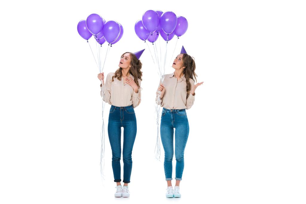 surpreso jovens gêmeos atraentes olhando para balões violetas isolados em branco
 - Foto, Imagem