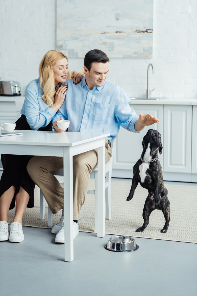 Man and woman drinking coffee and playing with their dog in kitchen - Photo, Image