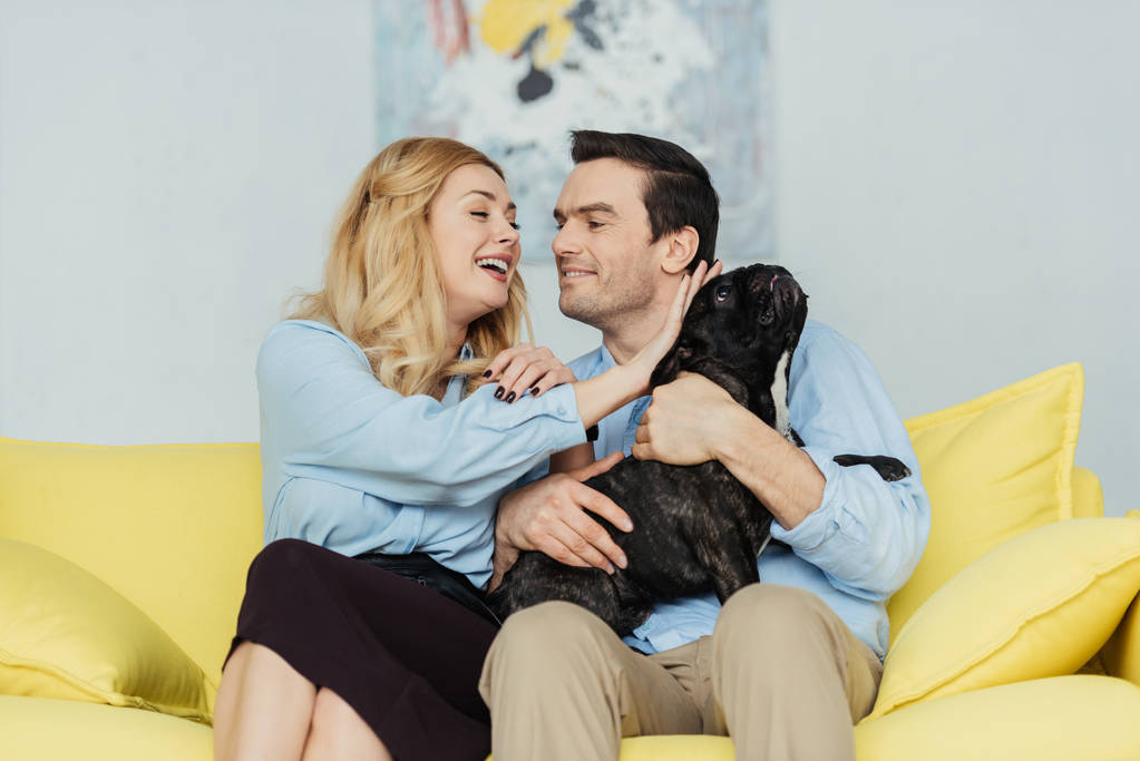 Para ładny patrząc na siebie nawzajem i przytulanie Frenchie psa na kanapie żółty - Zdjęcie, obraz