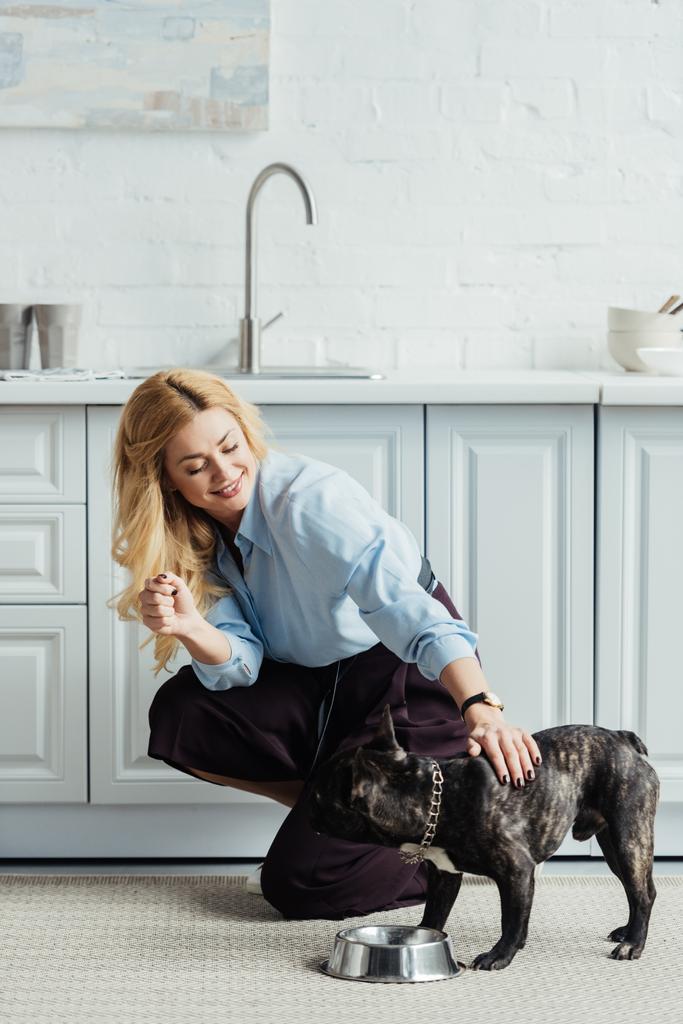 Sonriente mujer rubia acariciando frenchie perro en piso de cocina
 - Foto, imagen