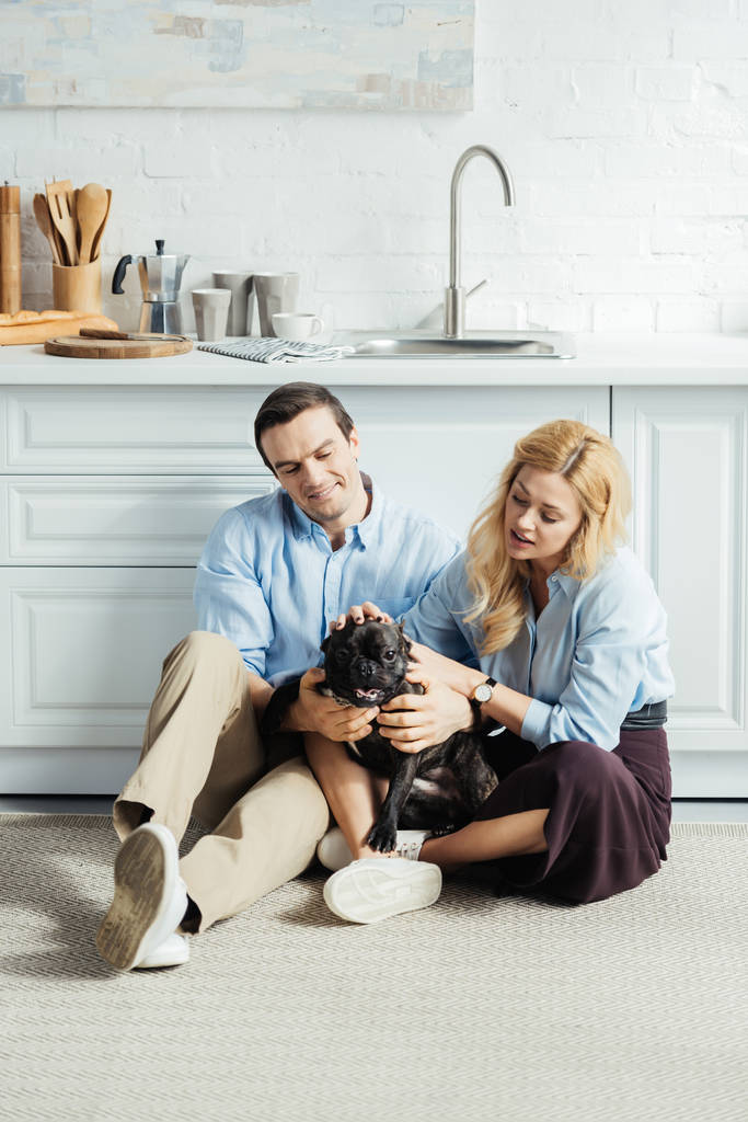 Feliz hombre y mujer rubia sentados en el suelo en la cocina con bulldog francés
 - Foto, imagen