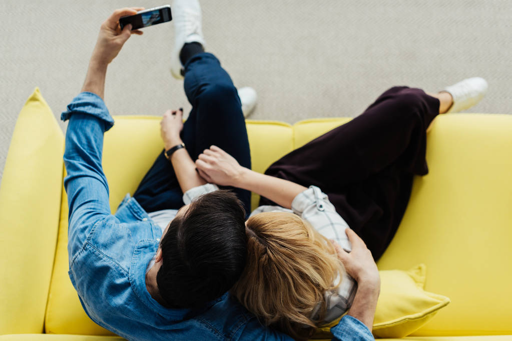 vista aerea di abbracciare uomo e donna seduti sul divano e scattare selfie
 - Foto, immagini