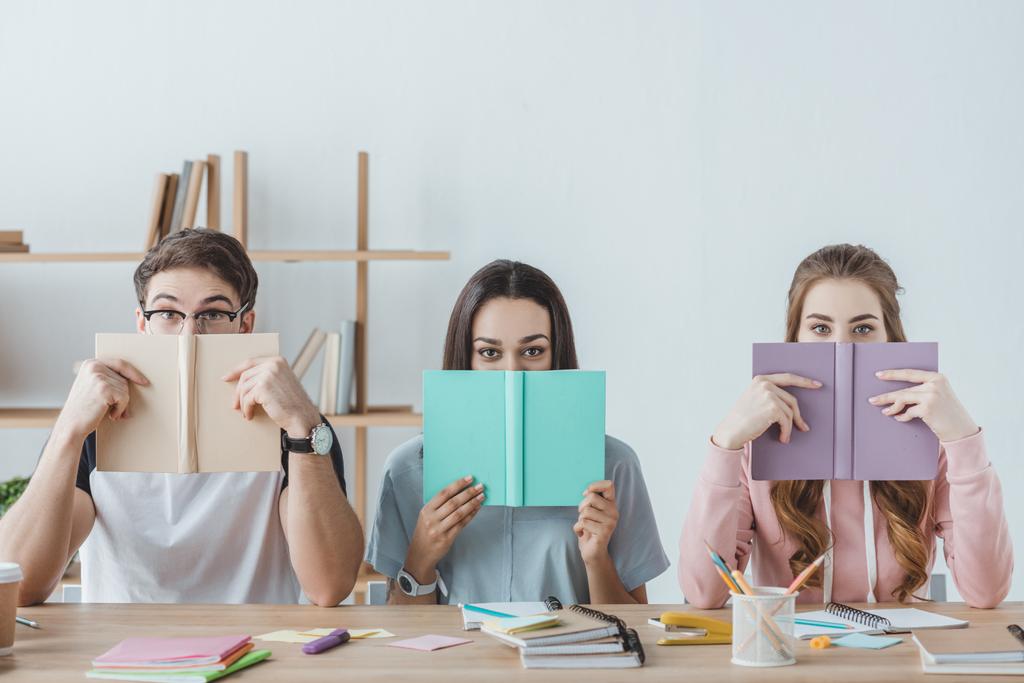 tres jóvenes estudiantes sosteniendo libros mientras están sentados en la mesa
  - Foto, imagen