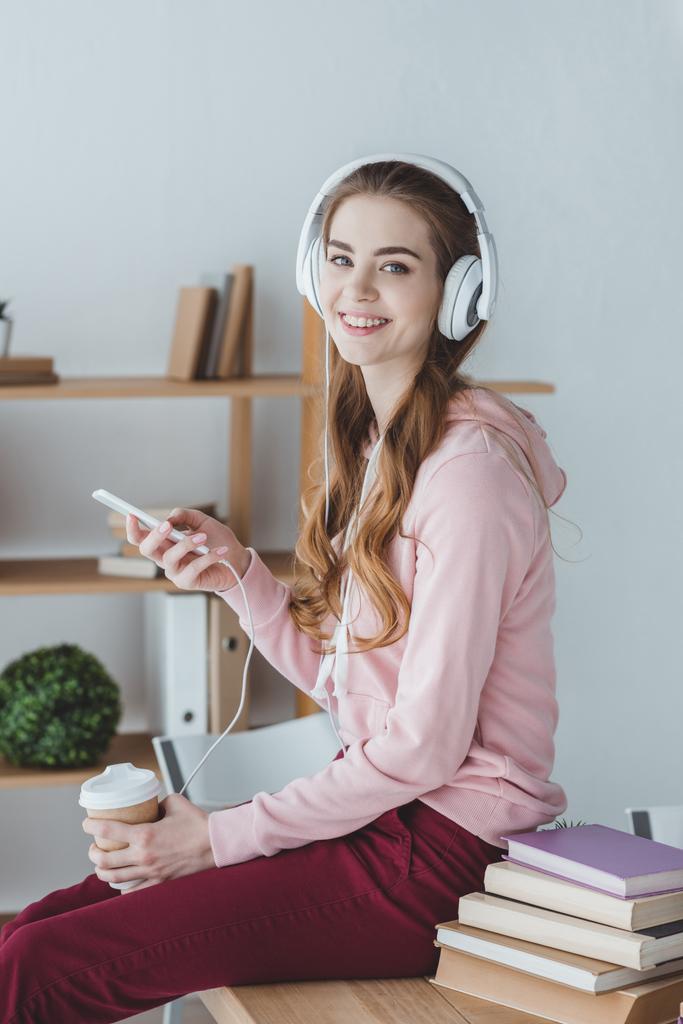 χαμογελώντας ξανθιά φοιτητής με καφέ ακούγοντας μουσική με το smartphone και ακουστικά - Φωτογραφία, εικόνα