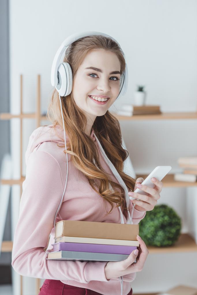 Lächelnde Studentin hält Bücher in der Hand und hört Musik mit Smartphone und Kopfhörer - Foto, Bild