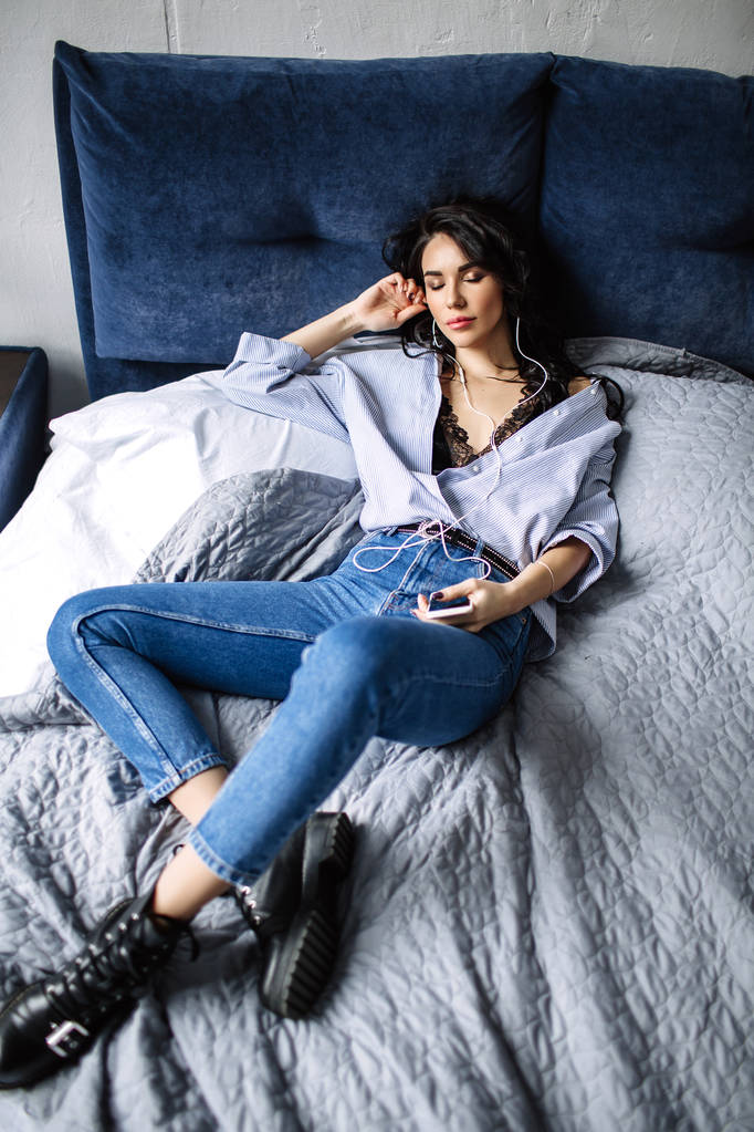 Piękna Brunetka słucha muzyki w słuchawkach w pokoju w pobliżu kanapie, jest ubrana w koszulę i niebieskie dżinsy, niebieski - Zdjęcie, obraz