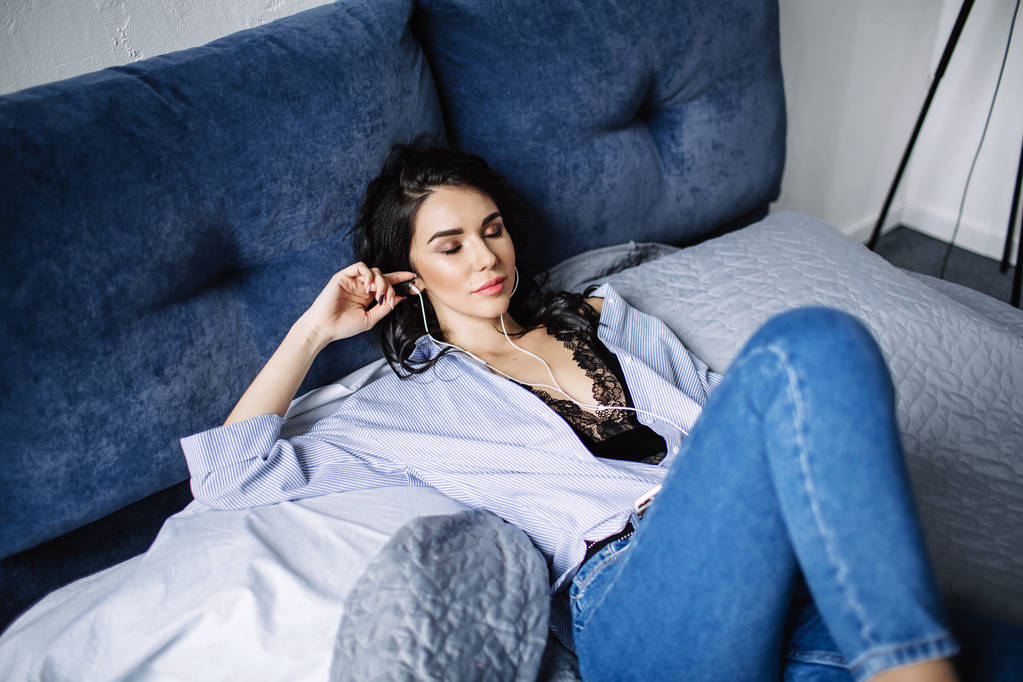 Piękna Brunetka słucha muzyki w słuchawkach w pokoju w pobliżu kanapie, jest ubrana w koszulę i niebieskie dżinsy, niebieski - Zdjęcie, obraz