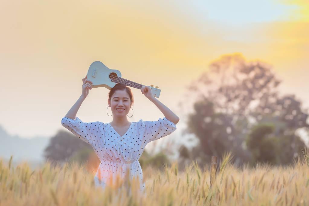 Hermosa mujer asiática sosteniendo ukelele y relajarse en el campo de cebada en verano en la hora de la puesta del sol
 - Foto, Imagen