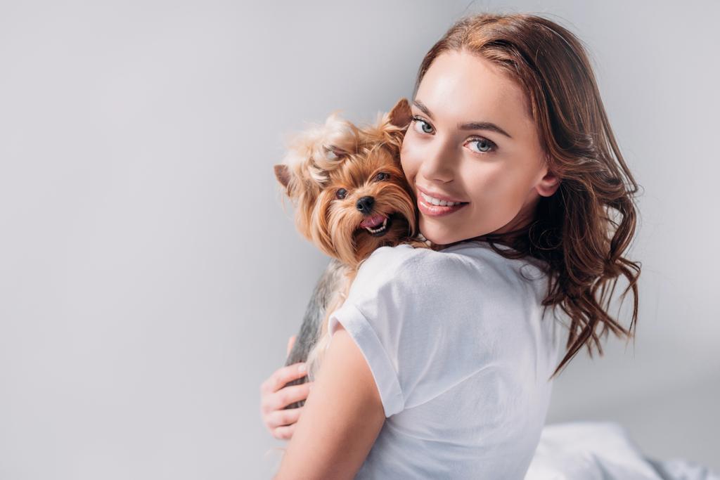Porträt einer jungen lächelnden Frau mit einem York Terrier, die isoliert auf grau in die Kamera blickt - Foto, Bild