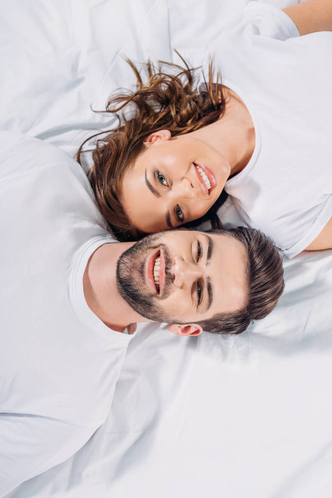 Вид сверху на молодую улыбающуюся влюбленную пару, которая смотрит в камеру, лежа вместе на кровати
 - Фото, изображение