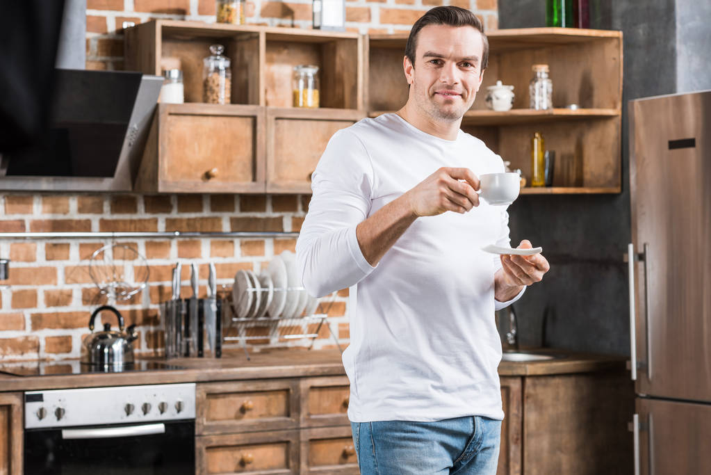 homem bonito segurando xícara de café e sorrindo para a câmera na cozinha
 - Foto, Imagem