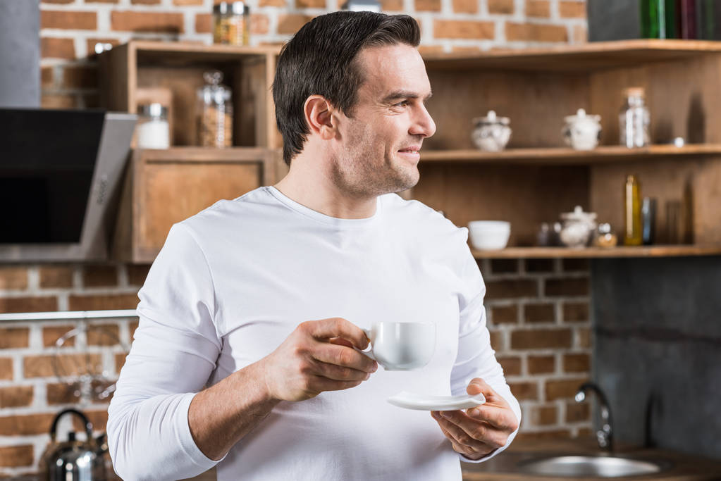 handsome man smiling and looking away while drinking coffee in kitchen - Photo, Image