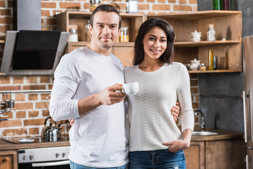 feliz pareja multiétnica sonriendo a la cámara mientras están de pie juntos en la cocina
 - Foto, imagen