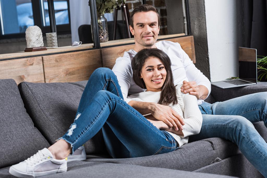 feliz pareja multiétnica sonriendo a la cámara mientras descansan juntos en el sofá en casa
 - Foto, imagen