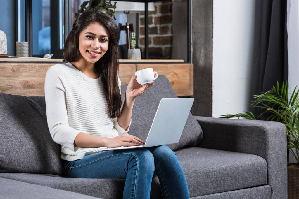 bella sorridente ragazza africana americana utilizzando il computer portatile e bere caffè sul divano
 - Foto, immagini