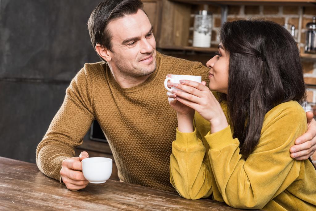 happy multiethnic couple drinking coffee and smiling each other at home - Photo, Image