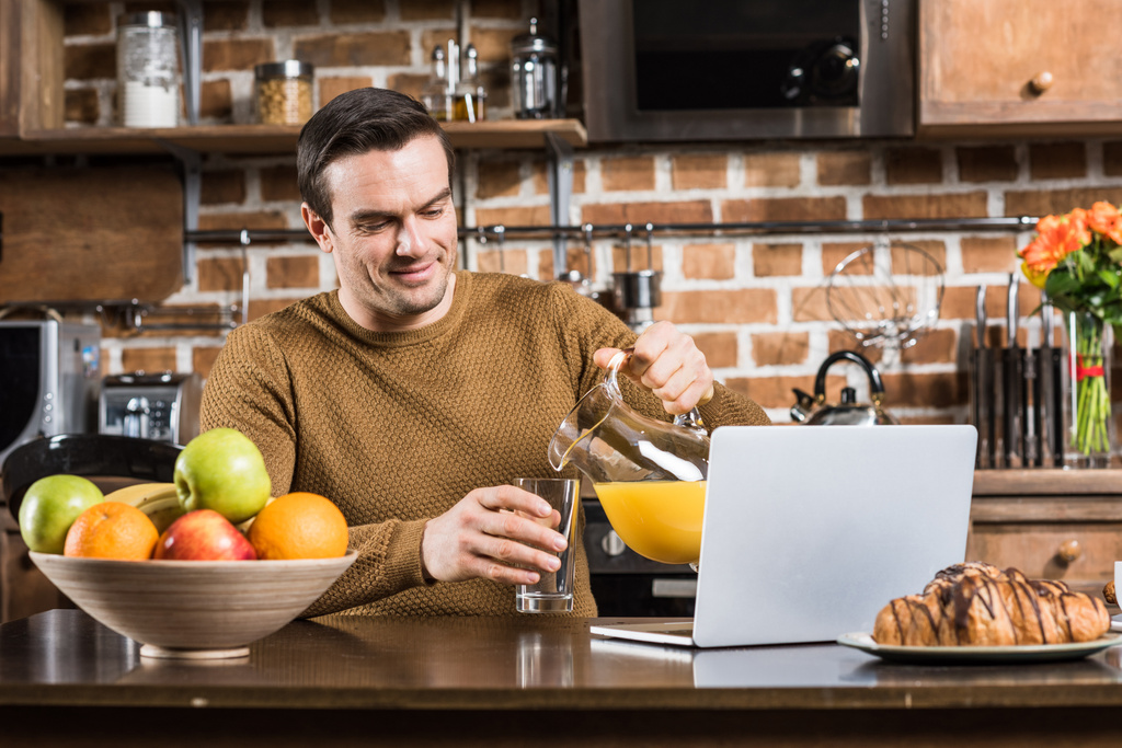 bonito sorrindo homem derramando suco enquanto usando laptop na mesa da cozinha
 - Foto, Imagem