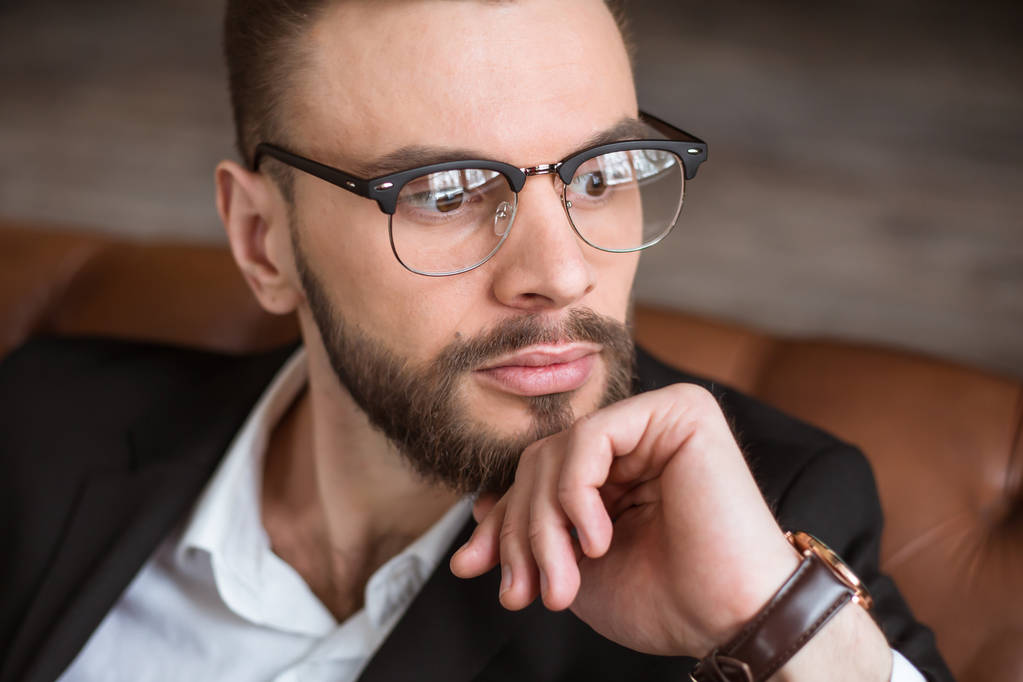 Όμορφος μοντέρνος αυτοπεποίθηση γενειοφόρος επιχειρηματία σε ένα έξυπνο κοστούμι και τα γυαλιά, κάθεται σε έναν καναπέ στο γραφείο. - Φωτογραφία, εικόνα