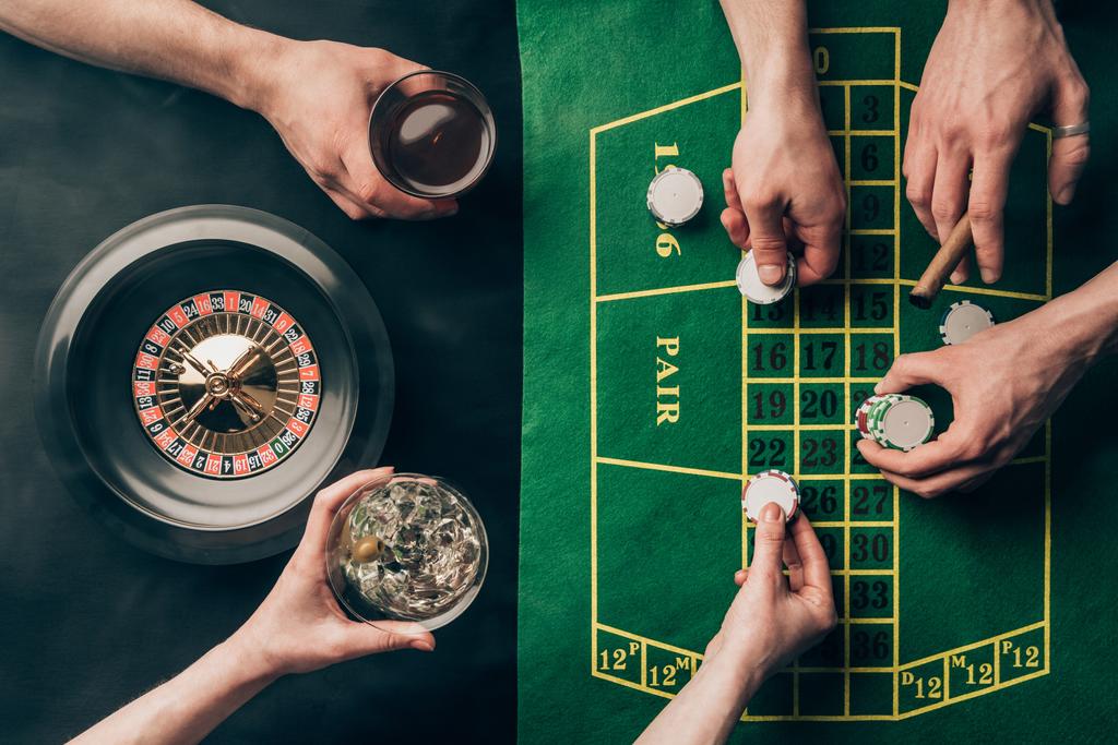 Χέρι με ουίσκι και τσιπ από τα τσιπ καζίνο στο τραπέζι με στοιχήματα και ρουλέτα - Φωτογραφία, εικόνα