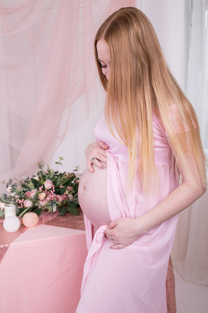 Acercamiento de la mujer embarazada tocándose el vientre y mirándola. Cuerpo de mujer durante el embarazo en vestido rosa sobre fondo con flor. Sus manos en el estómago, las manos sobre la barriga. Esperanza
 - Foto, Imagen