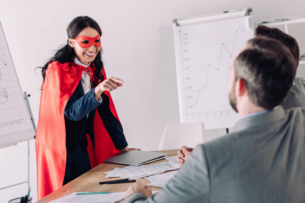 smiling super businesswoman in mask and cape pointing on businessmen in office - Photo, Image