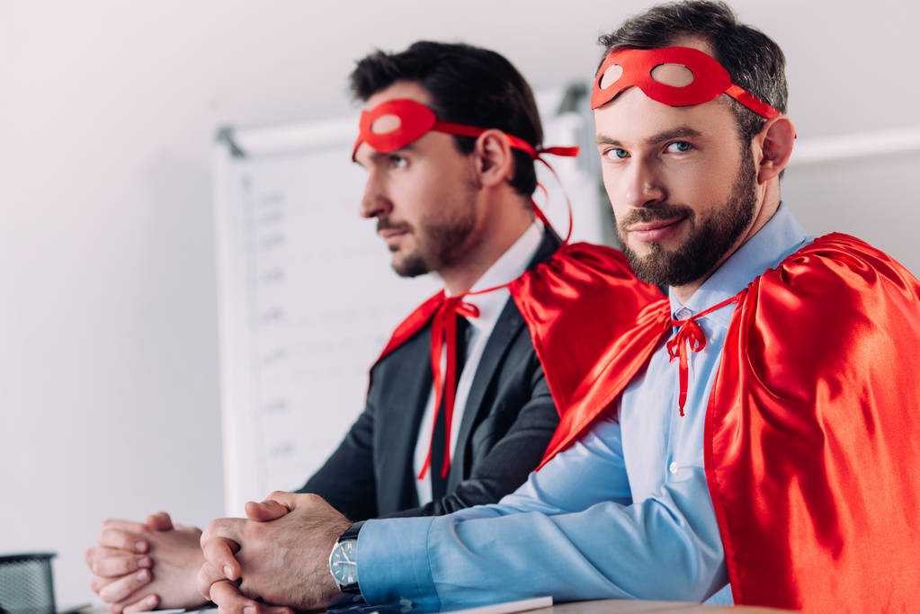 knappe super zakenlieden in maskers en capes zittend aan tafel in kantoor - Foto, afbeelding