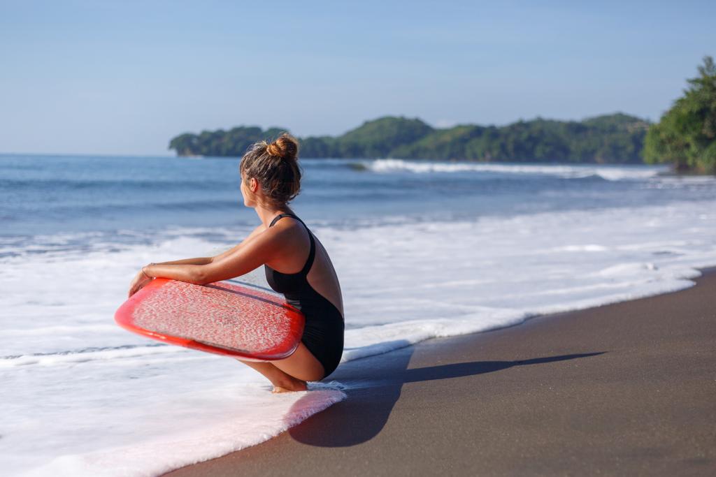 молодой серфер в купальнике сидит с красной доской для серфинга у моря
 - Фото, изображение