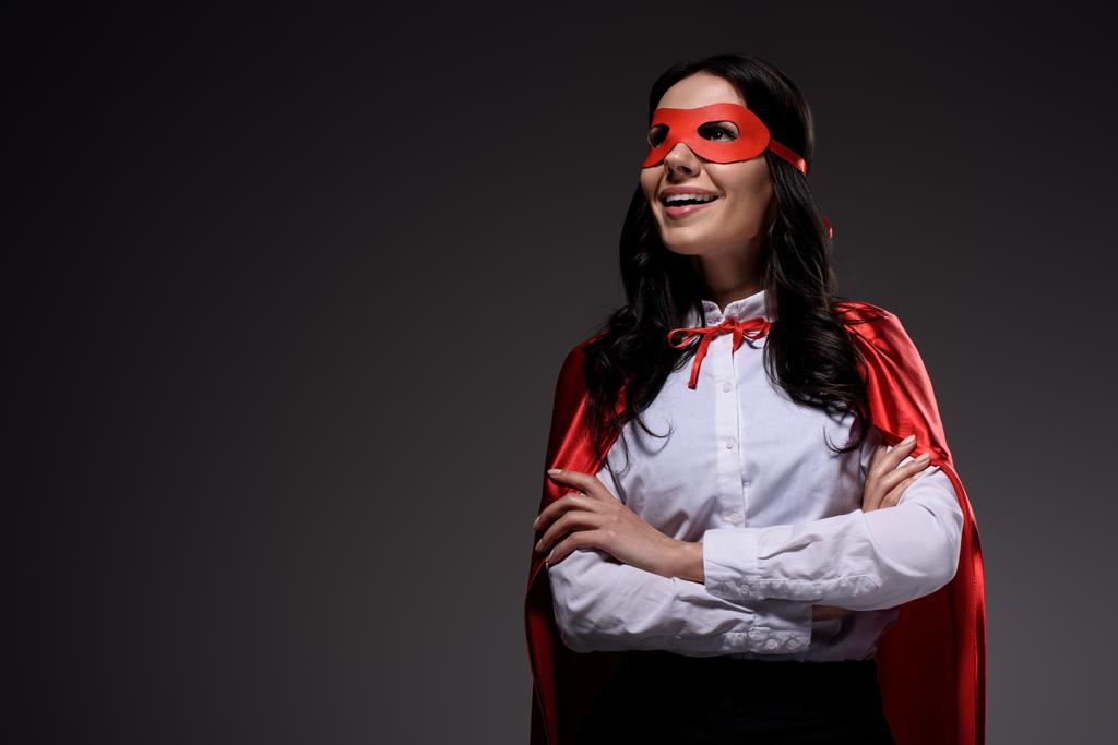 smiling attractive super businesswoman in red cape and mask standing with crossed arms isolated on black - Photo, Image