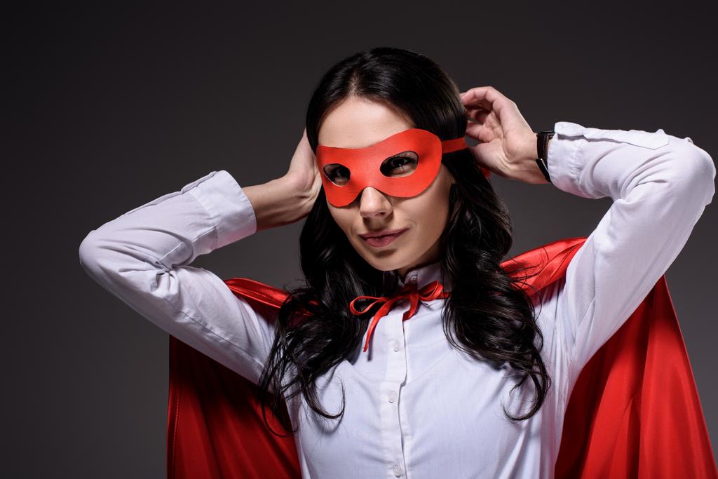 ελκυστική σούπερ επιχειρηματίας στην κόκκινη κάπα δένοντας μάσκα που απομονώνονται σε μαύρο - Φωτογραφία, εικόνα