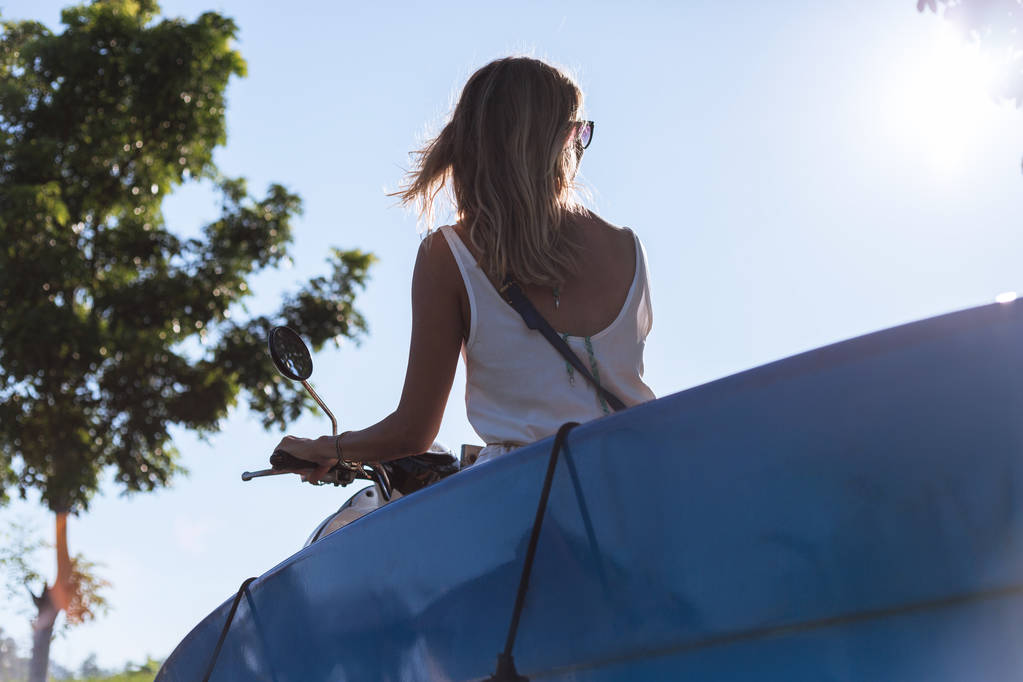 青い空を背景にサーフィン ボードを持つ女性乗馬スクーターの背面図 - 写真・画像