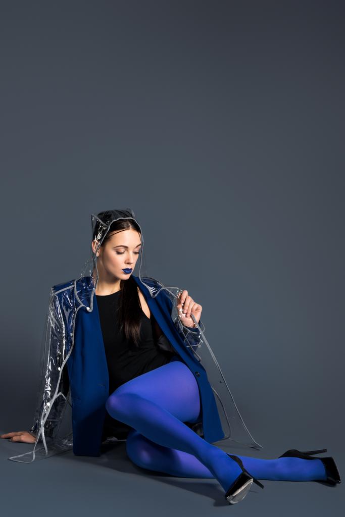 Tyttö sinisissä vaatteissa ja läpinäkyvä sadetakki makaa tummalla taustalla
 - Valokuva, kuva