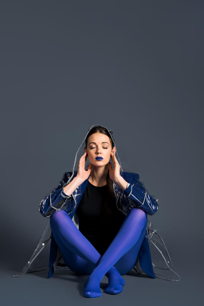 Девушка в прозрачном плаще и синих колготках сидит на тёмном фоне
 - Фото, изображение