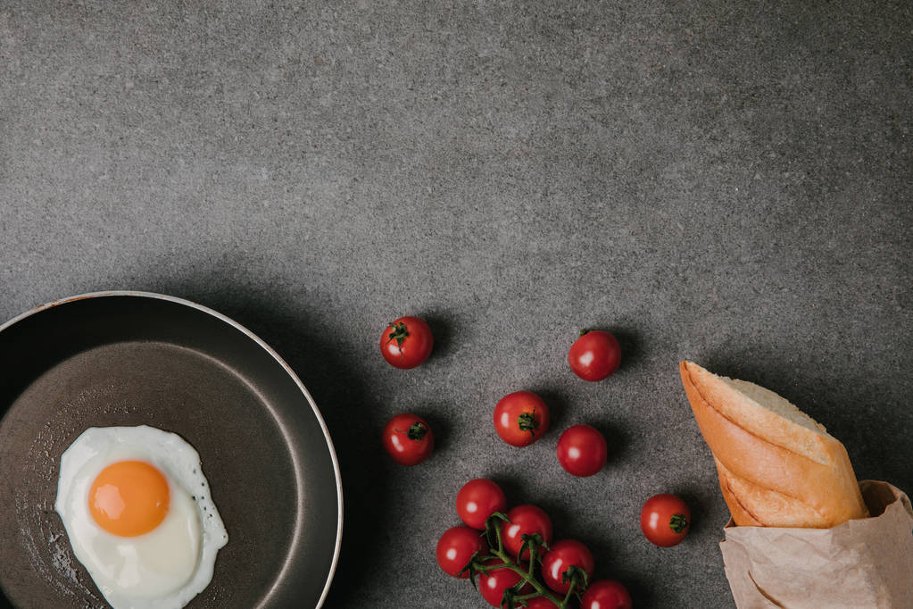 верхний вид жареного яйца на сковороде, свежие помидоры и багет в бумажном пакете на сером
 - Фото, изображение