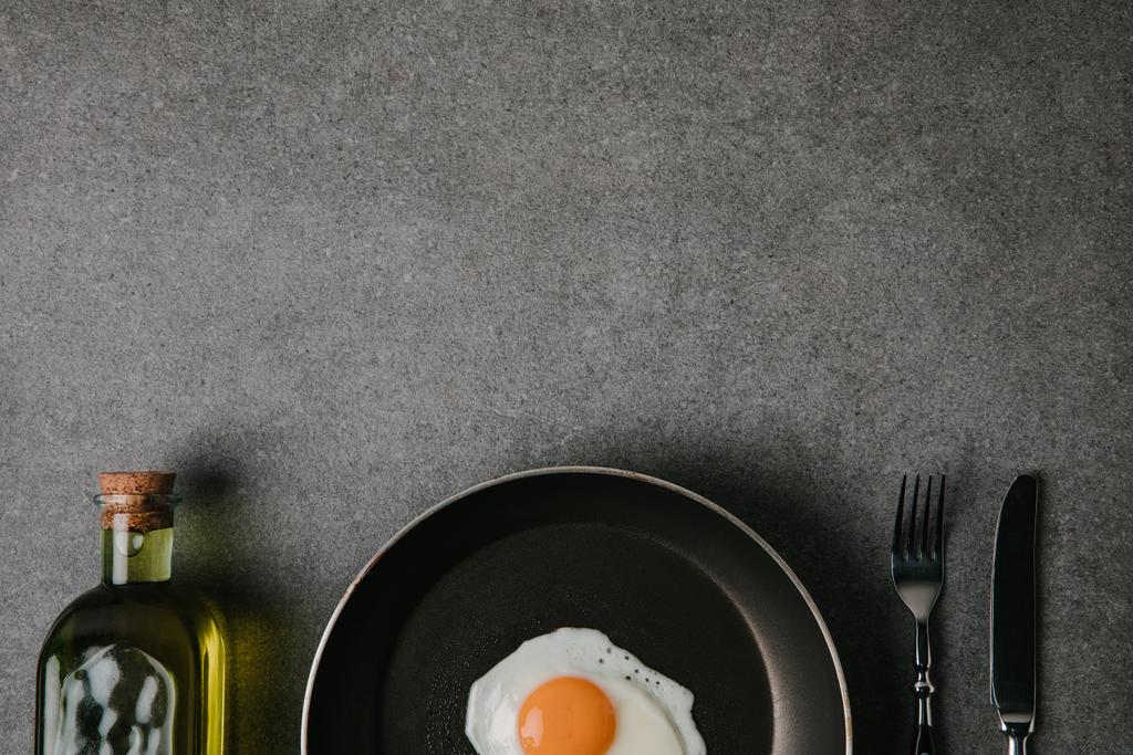 верхний вид сковороды с жареным яйцом, столовые приборы и бутылка масла на сером
  - Фото, изображение
