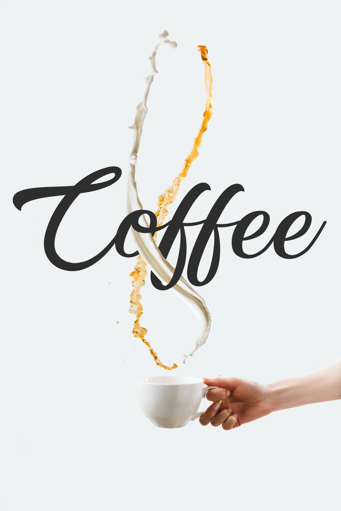 cropped view of hand holding cup with splashes of milk and hot coffee, isolated on white with "Coffee" symbol - Photo, Image