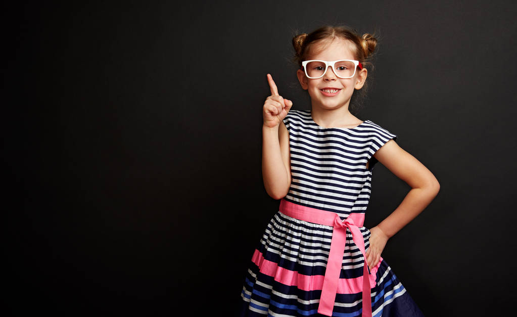 Menina muito sorridente em óculos apontando sobre fundo preto. Criança alegre em vestido elegante com ideia criativa
. - Foto, Imagem